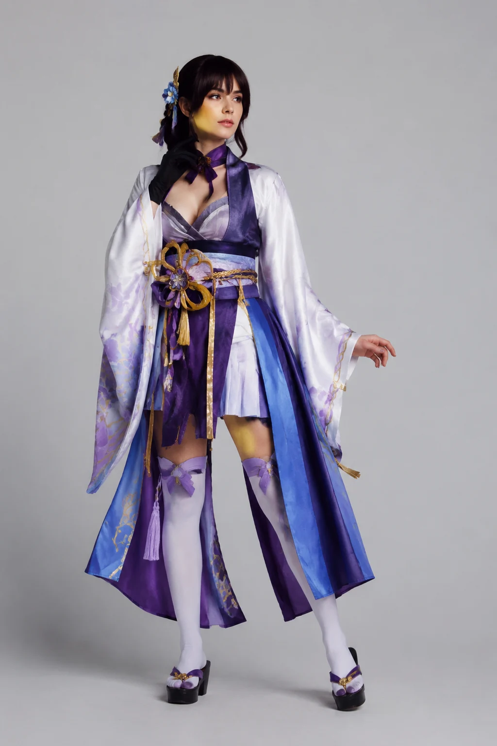 A person in a colorful anime-inspired cosplay with layered fabric and a decorative belt, posed against a plain light-gray studio background in soft light