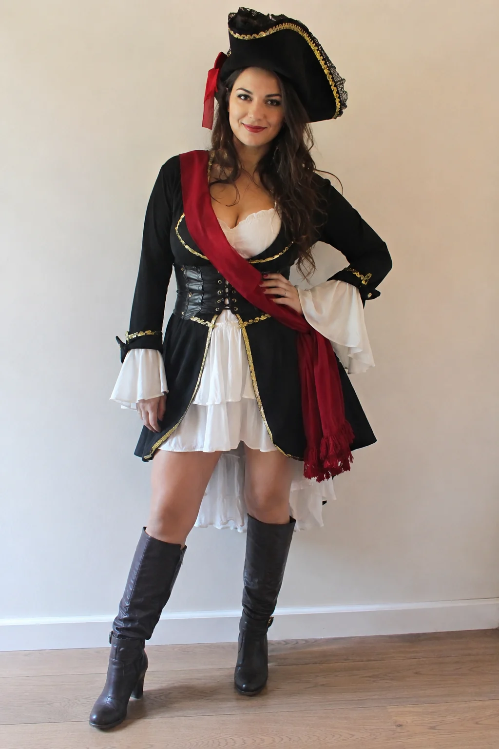 A woman in a playful pirate-themed party costume with a sash and hat, photographed full-length in even light against a plain wall