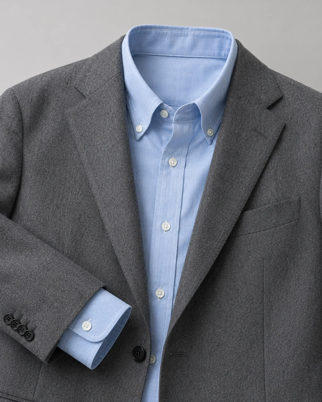 Product shot of a light blue oxford shirt and a grey wool blazer on a plain neutral background, crisp editorial lighting