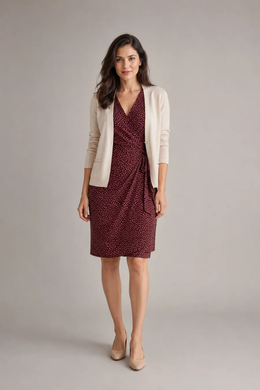 A woman in her late twenties wearing a burgundy knee-length wrap dress with a small white dot print, a cream tailored cardigan, and tan low heels, standing upright facing forward, full body, entire figure from head to feet in frame, centered with headroom