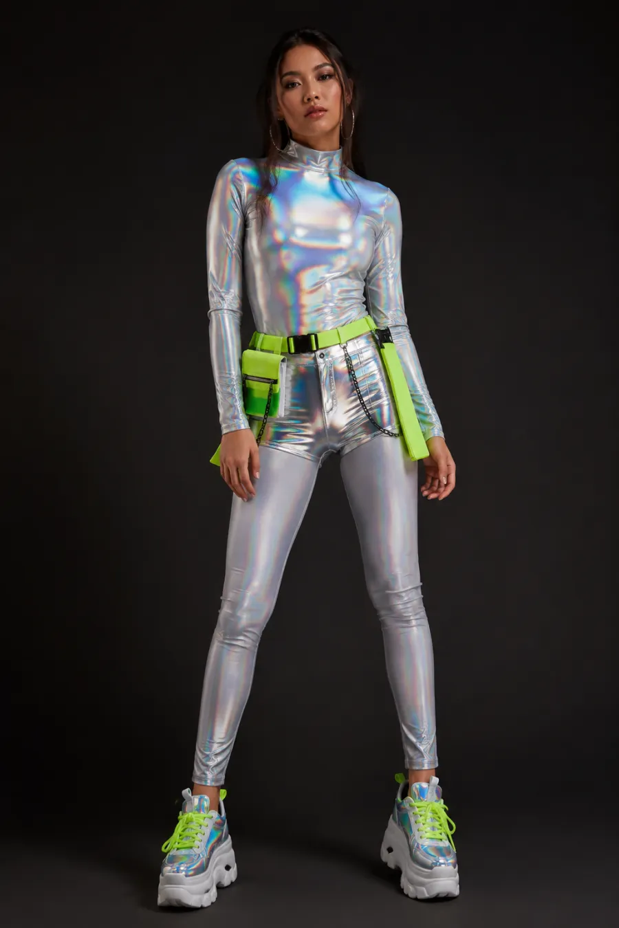 Rave festival outfit: a holographic long-sleeve top, metallic high-waisted shorts, a neon utility belt, and chunky platform sneakers shown on a model against a plain dark studio background, vivid lighting, full body