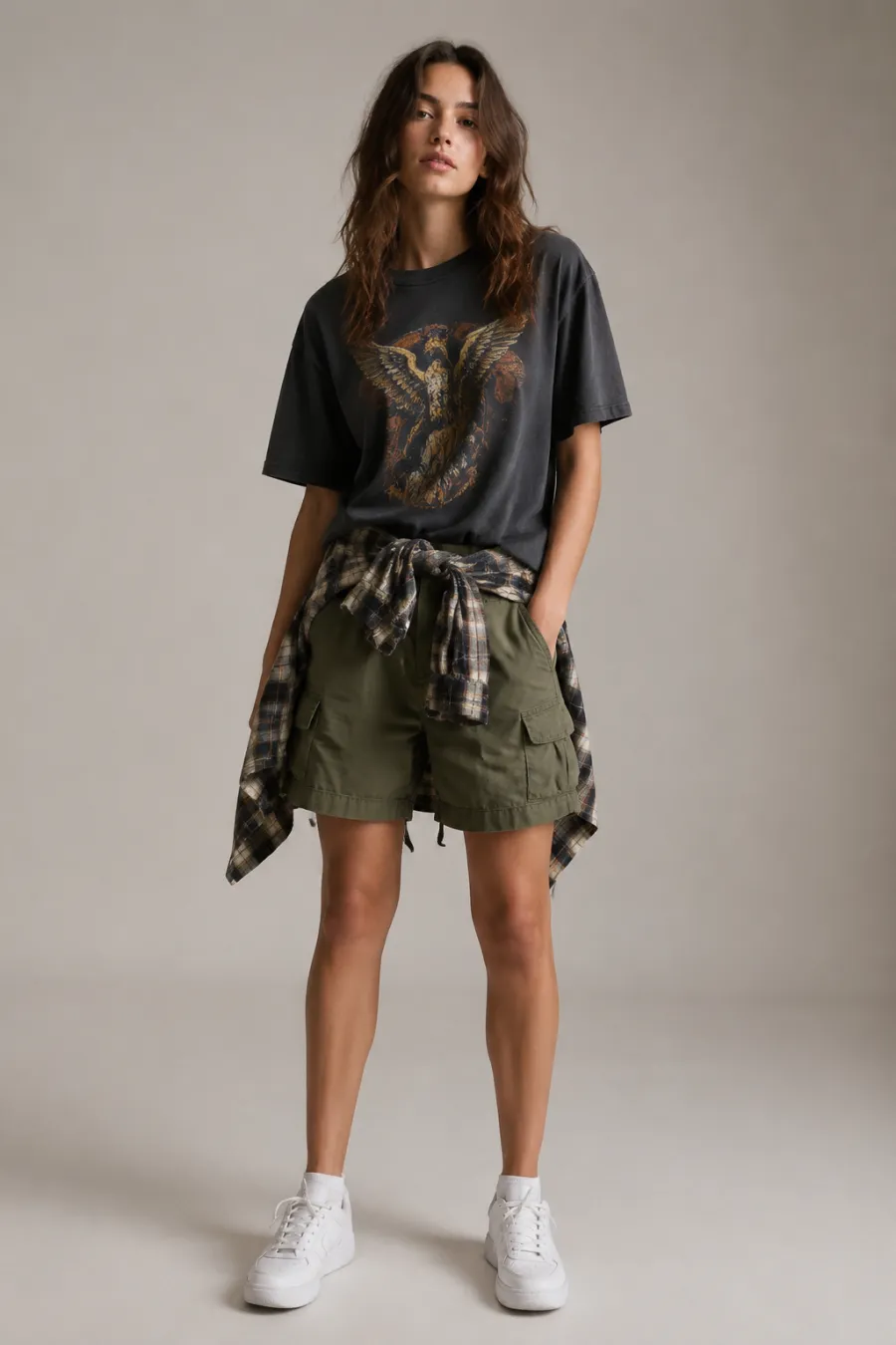 Casual unisex festival outfit: vintage graphic tee, olive cargo shorts, flannel shirt tied at the waist, and white sneakers shown on a model against a plain neutral background, clean studio lighting, full body