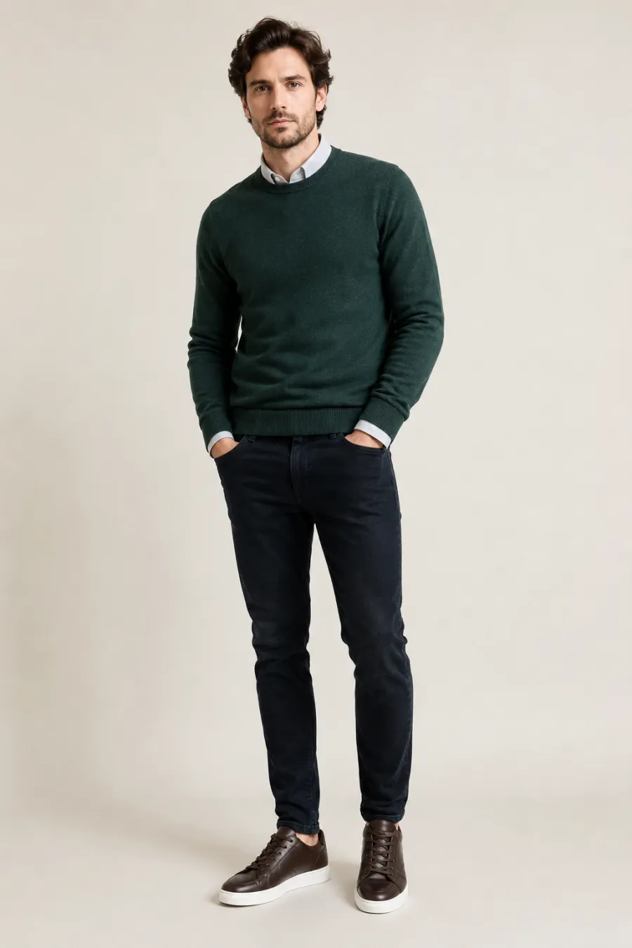A smart-casual creative interview look — a dark green fine-knit sweater over a collared shirt with dark slim jeans and clean leather sneakers, shown on a plain light background, full body