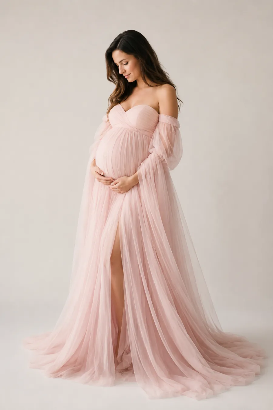 Full-body shot of a flowing blush-pink tulle empire-waist maternity dress with a long trailing skirt, shown on a plain light background with airy soft lighting