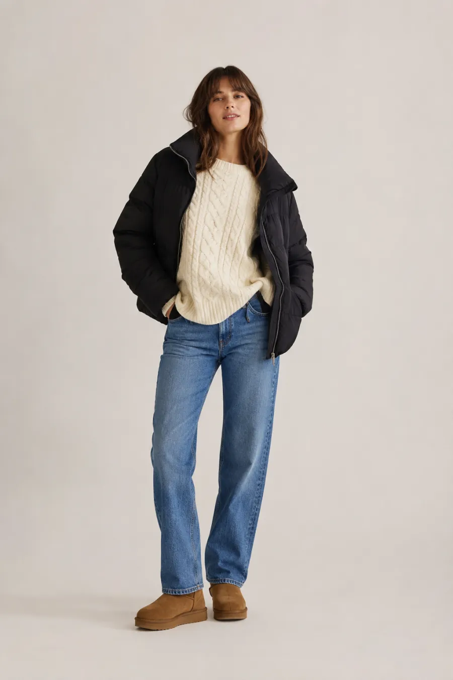 Full body shot of a casual winter outfit on a woman in her twenties — an oversized cream cable-knit sweater, blue straight-leg jeans, a black puffer jacket, and tan ankle boots, on a plain light studio background