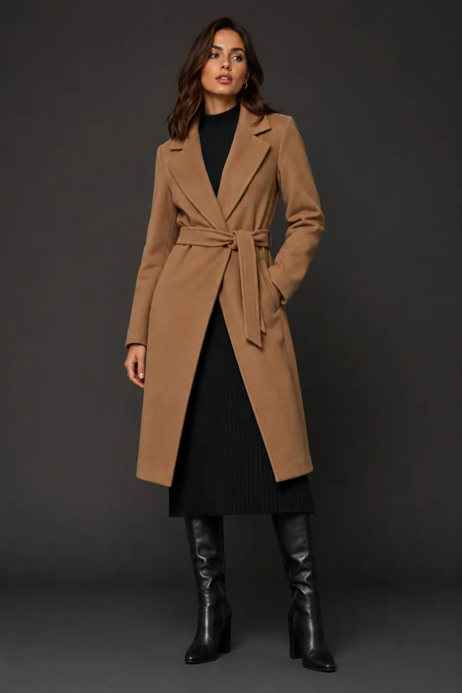 Full body shot of a dressy winter outfit on a woman in her late twenties — a belted camel wrap coat over a black ribbed knit midi dress with tall black heeled boots, on a plain dark studio background