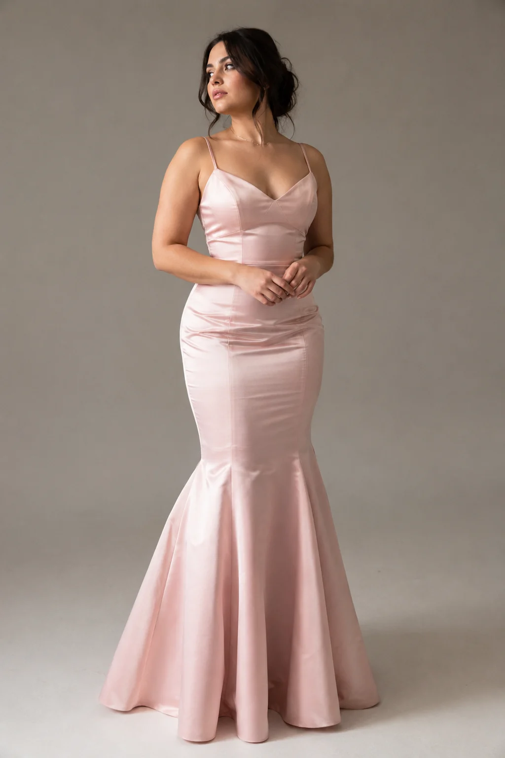 A young woman in a blush pink mermaid prom gown, fitted through the hips and flaring at the knee, photographed on a plain studio background in soft natural light