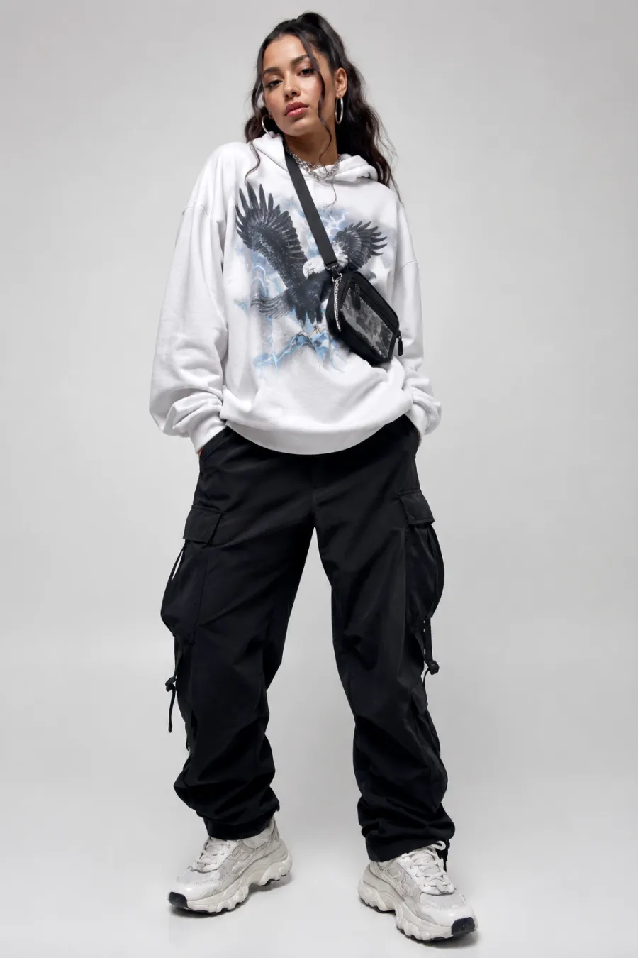 Rap concert streetwear outfit: a young woman in an oversized white graphic hoodie, baggy black cargo pants, chunky white sneakers, and a small crossbody bag, urban concert backdrop, full body