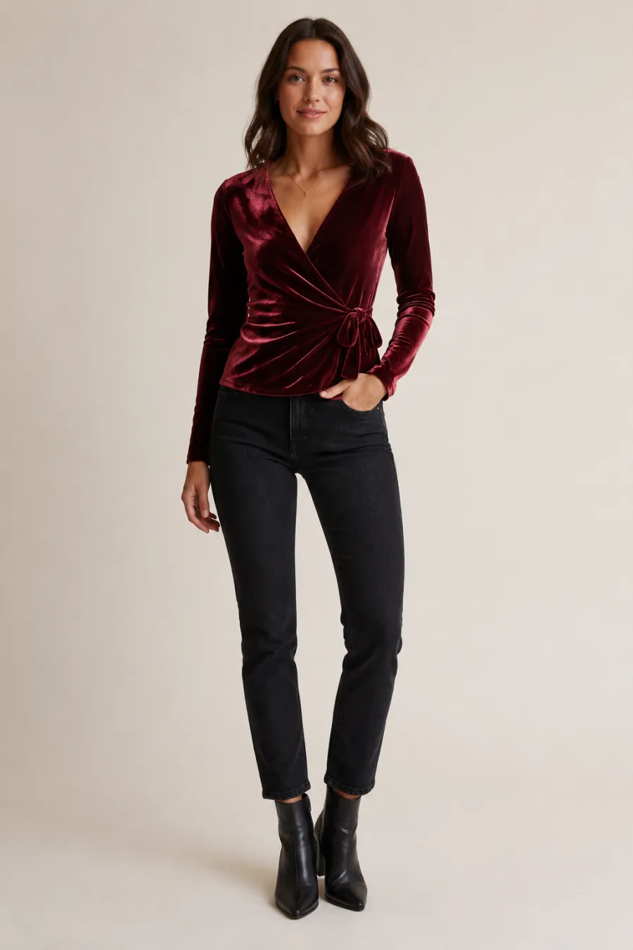 A burgundy velvet wrap top with dark jeans and ankle boots for a cozy New Year's Eve at home, shown on a plain light studio background with soft warm lighting, full body