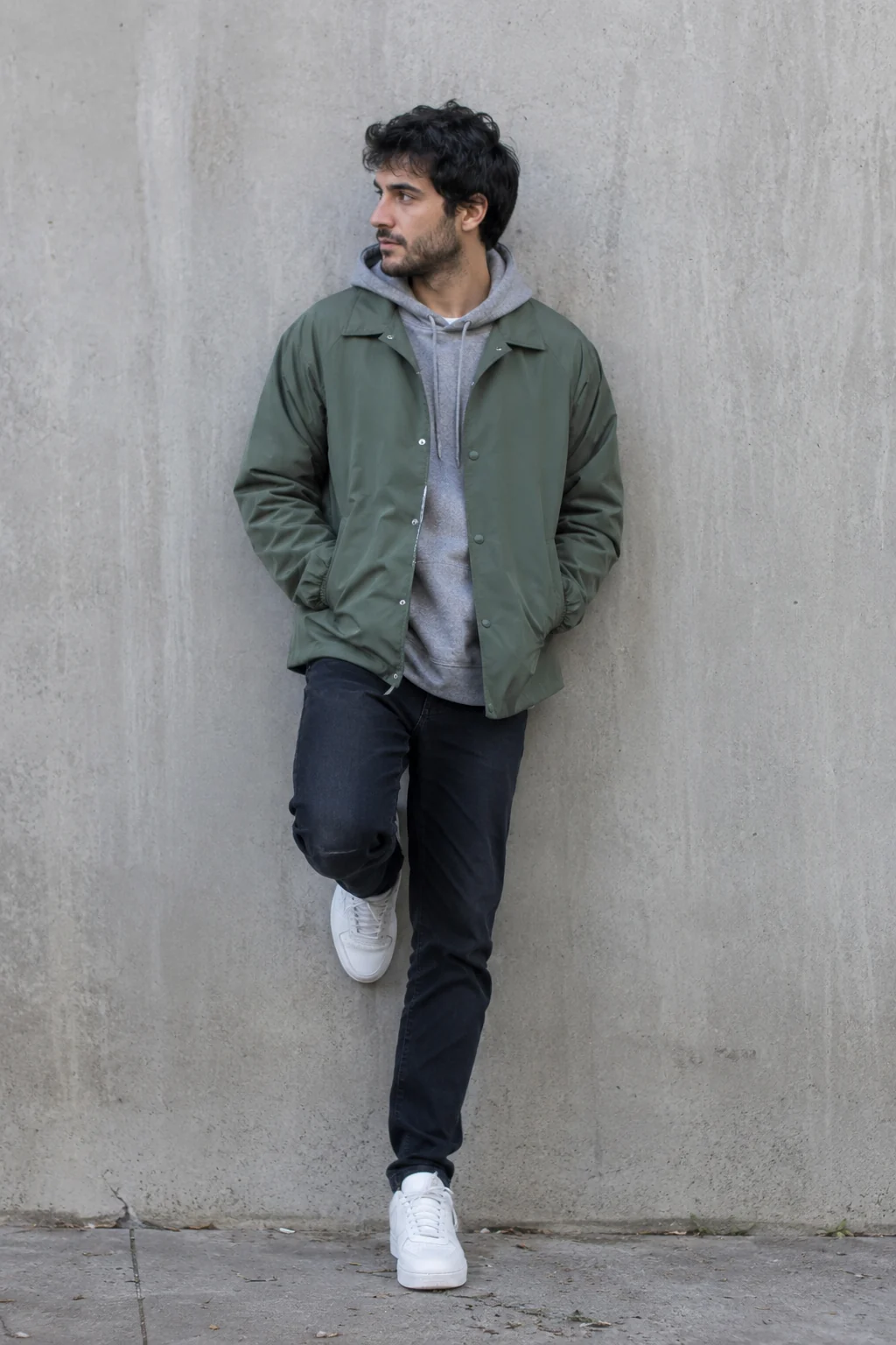 A man in his late twenties standing against a concrete wall in a green coach jacket over a grey hoodie, dark jeans and white sneakers, natural daylight