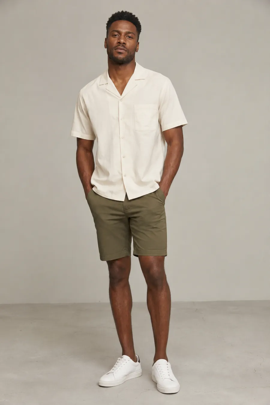 An adult Black man wearing a cream short-sleeve camp-collar shirt and olive cotton chino shorts with white leather sneakers, relaxed daytime stance, full body, entire figure from head to feet in frame, centered with headroom