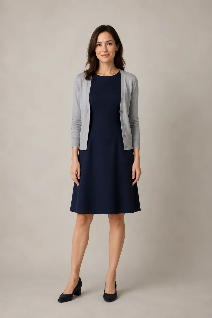 A woman in her early thirties wearing a navy knee-length A-line dress in matte crepe with a soft gray cardigan and low block heels, hands relaxed at her sides, full body, entire figure from head to feet in frame, centered with headroom