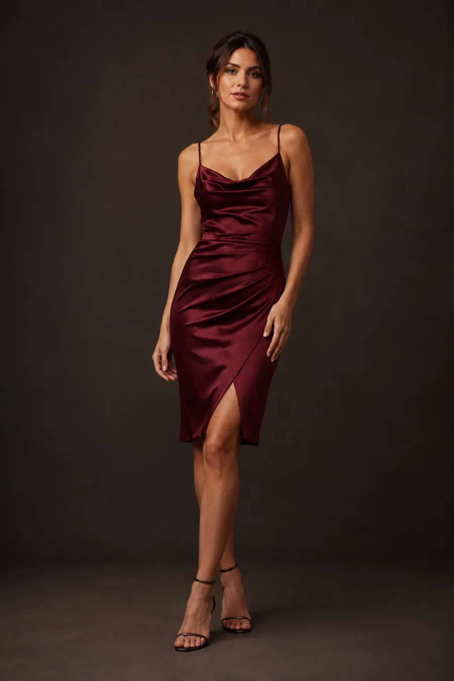 A woman in her late twenties wearing a knee-length burgundy satin cocktail dress with strappy heels, standing in soft evening light against a plain dark backdrop, full body