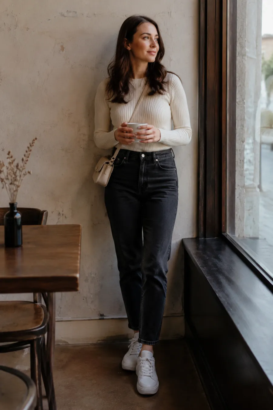 A woman in her mid twenties on a casual first date wearing high-waisted dark jeans, a fitted cream sweater and white sneakers with a small crossbody bag, standing in a cozy coffee shop by a window, full body