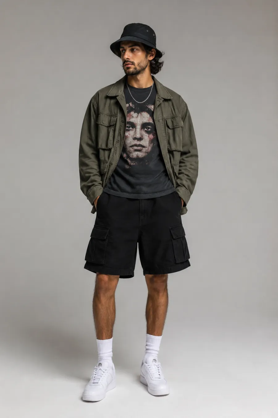A man in his late twenties at an outdoor festival at dusk wearing a graphic tee, an open olive utility jacket, black cargo shorts, and white sneakers, with a bucket hat, string lights and a crowd softly blurred behind him, full body