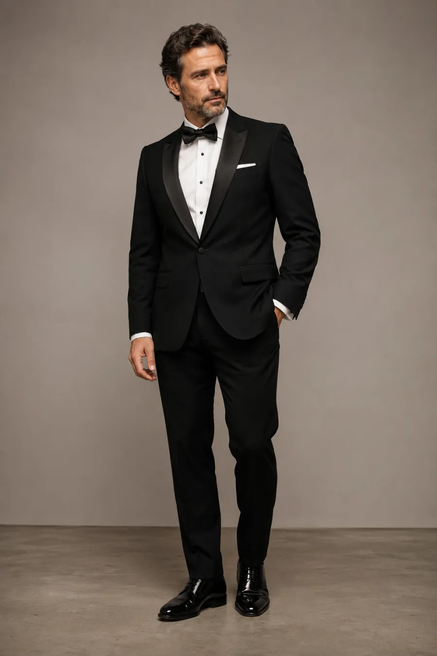 A man in his forties in a classic black tuxedo with satin lapels, black bow tie, and patent shoes, standing at an elegant evening gala reception, full body