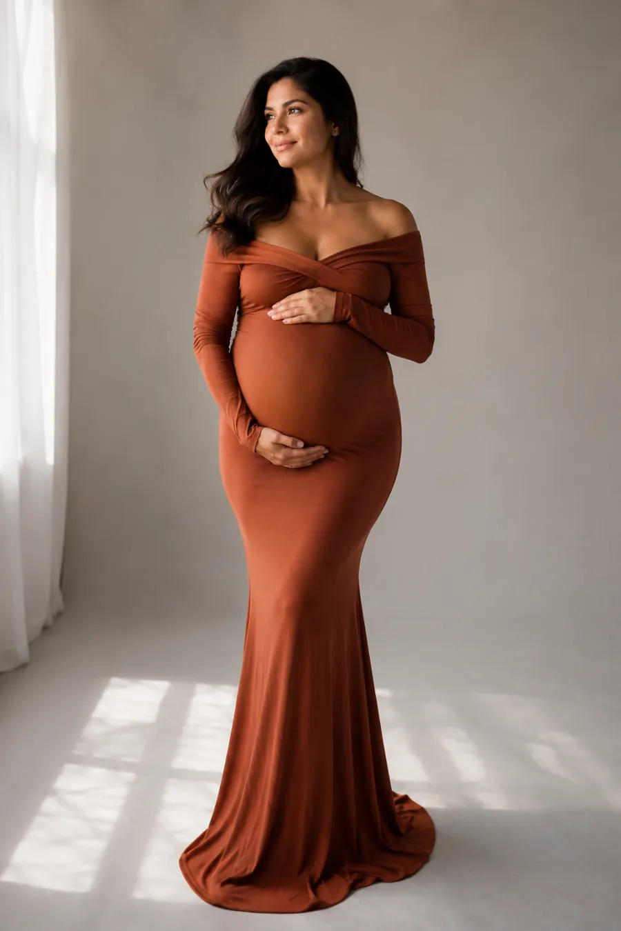 A pregnant woman in her late twenties wearing a fitted floor-length terracotta bodycon maternity gown that hugs her bump, standing in soft window light against a plain neutral backdrop, full body