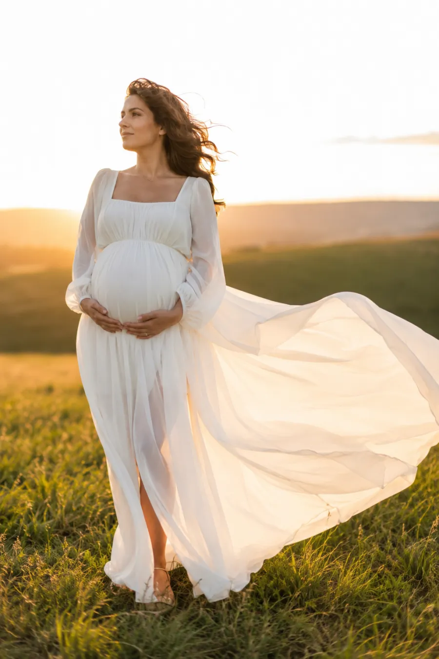 A pregnant woman in her thirties wearing a flowing white chiffon empire-waist maternity dress with long sleeves, fabric billowing softly in a breeze, photographed in golden-hour light in an open grassy field, full body