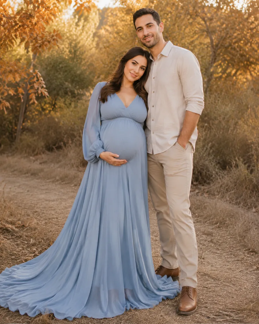 A pregnant woman in a long dusty-blue flowing maternity gown standing beside her partner in a matching neutral shirt and trousers, both in coordinated earth tones, outdoor autumn setting with warm light, full body
