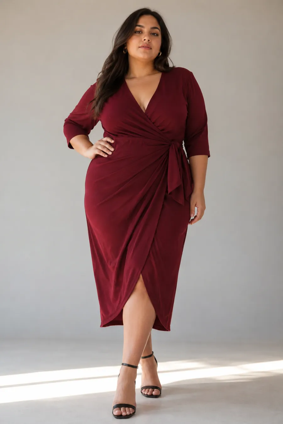 A confident plus size woman in her late twenties wearing a deep burgundy wrap dress with a defined waist, standing in soft daylight against a plain studio backdrop, full body