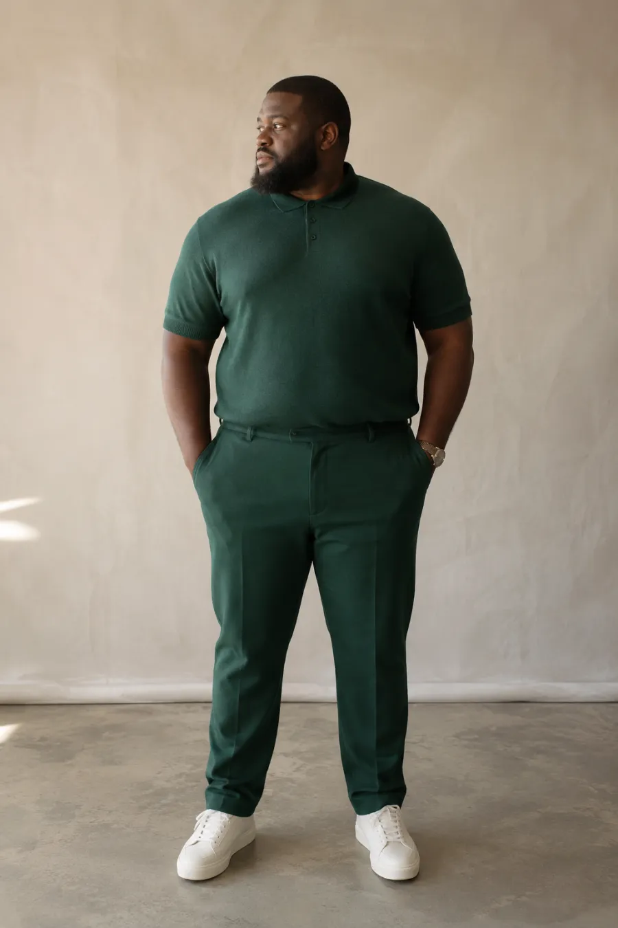 A plus size man in his thirties in a tailored forest-green monochrome outfit, knit polo and matching trousers, photographed in natural light against a neutral wall, full body