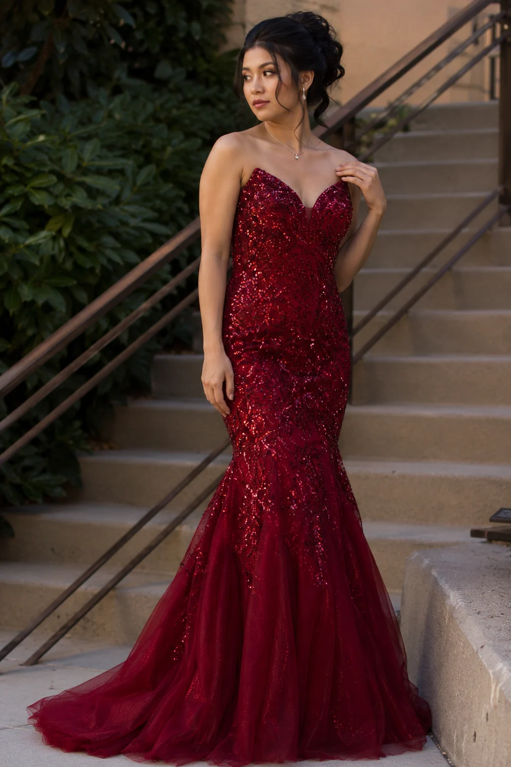 Close-up of a young woman in a deep ruby sequin mermaid prom dress, the beading catching light along the bodice, fabric flaring at the knee, natural editorial lighting