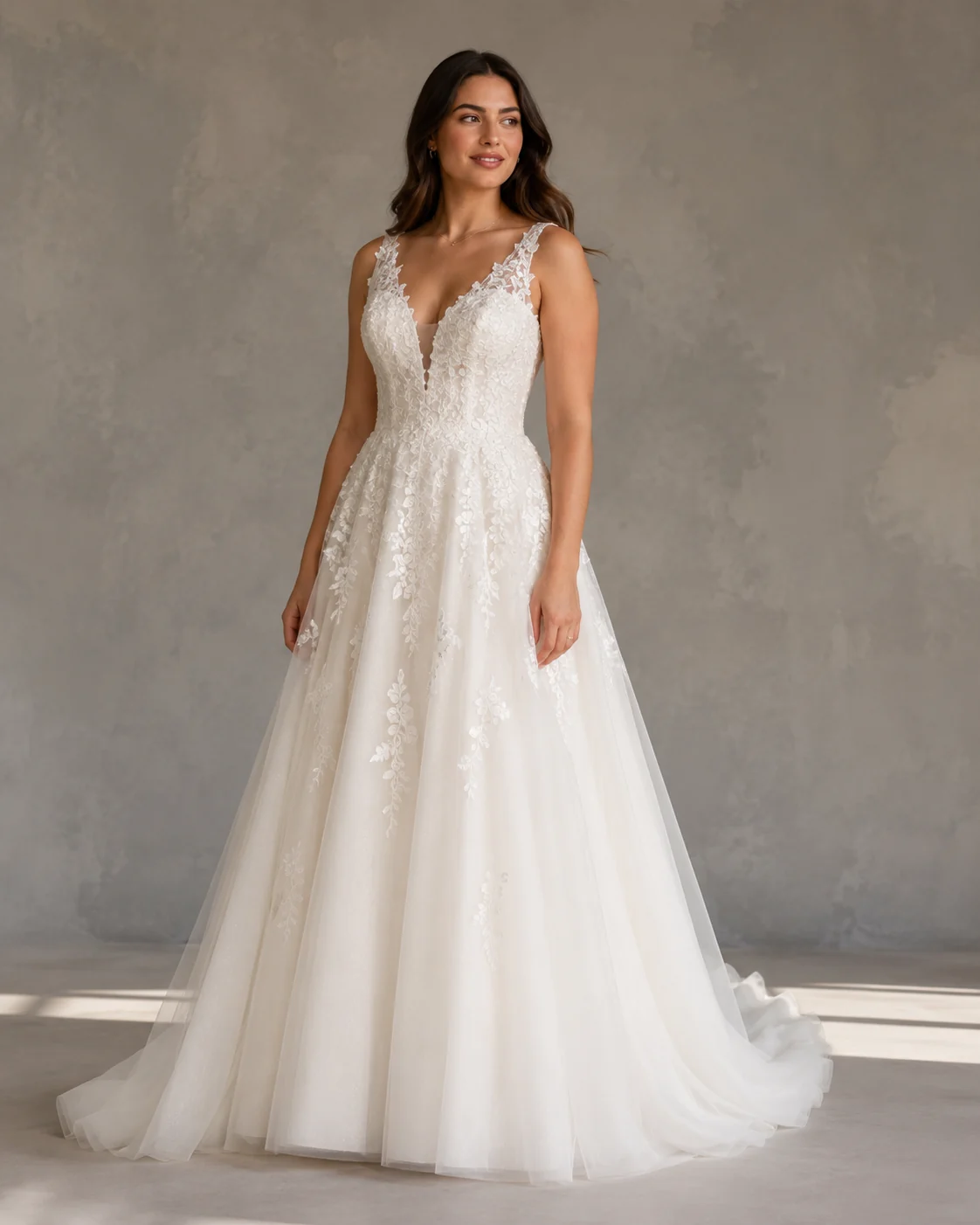 A bride-to-be in her twenties standing in soft daylight wearing an A-line ivory wedding gown, hands resting at her sides, plain studio backdrop